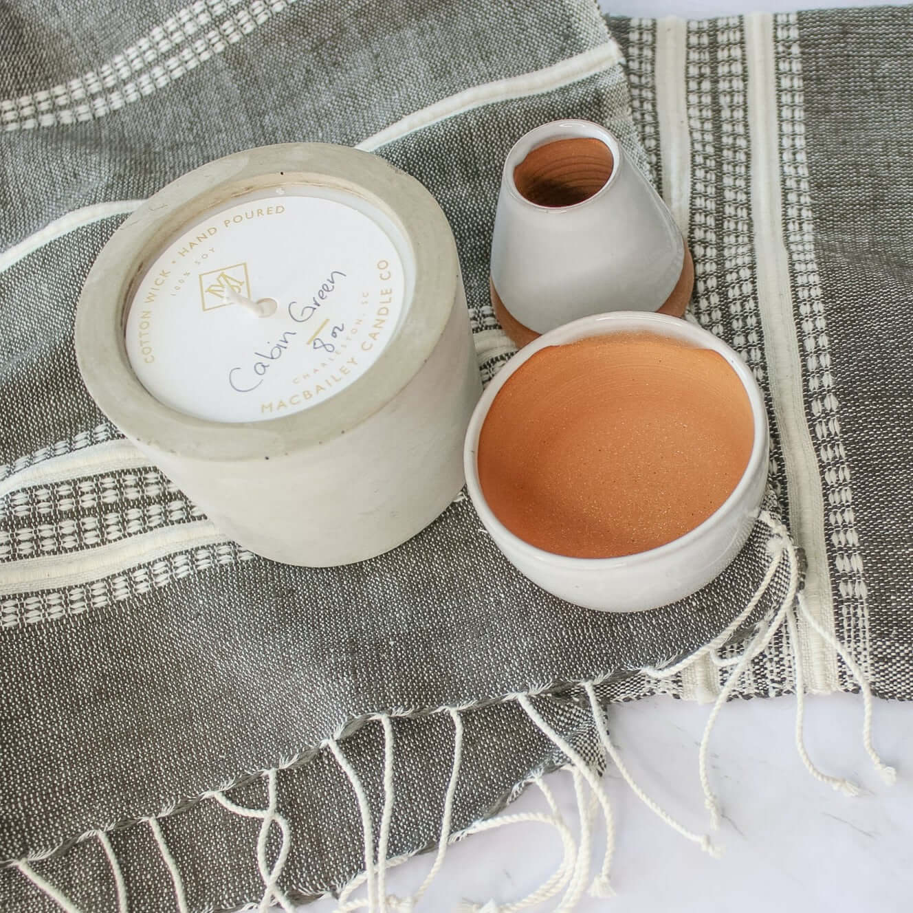 8 oz concrete candle resting on our grey hatch tea towel with tassels. Two of our ceramic planters are sitting next to the candle. All resting on a marble countertop.