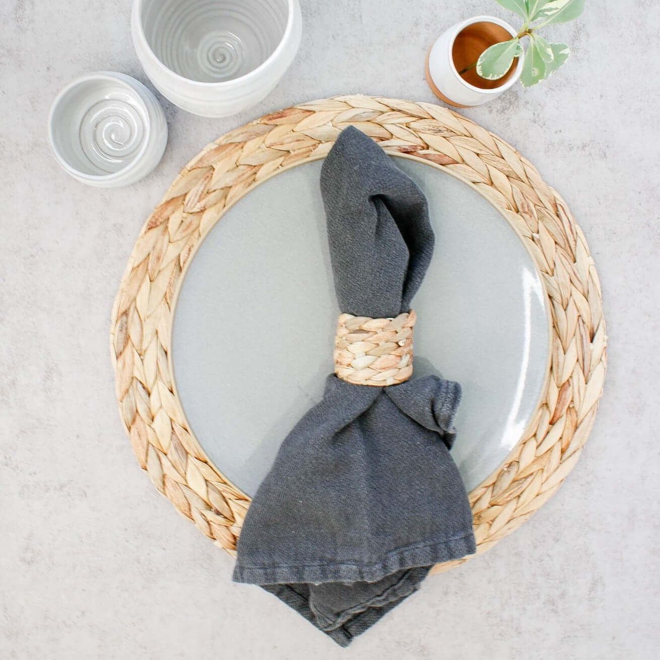A table place setting using our Seagrass placemat, cloth napkin in Abyss and seagrass napkin ring. Next to this is our Tall Tumbler, Tipsy Tumbler and on of the Tiny Planters.