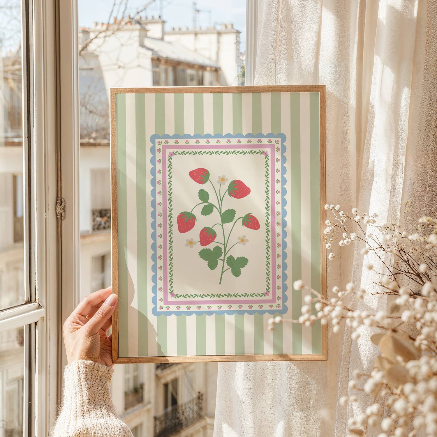 Framed artwork with floral and strawberry design held by a person in front of a window