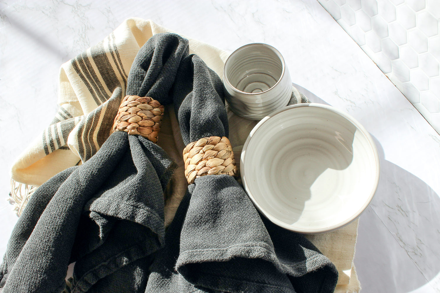 In beautiful morning light, our hatch cotton tea towel lays on a marble countertop. Two cloth napkins with our seagrass napkin rings sit on top of the towel. Our favorite bowl and tipsy tumbler complete the display