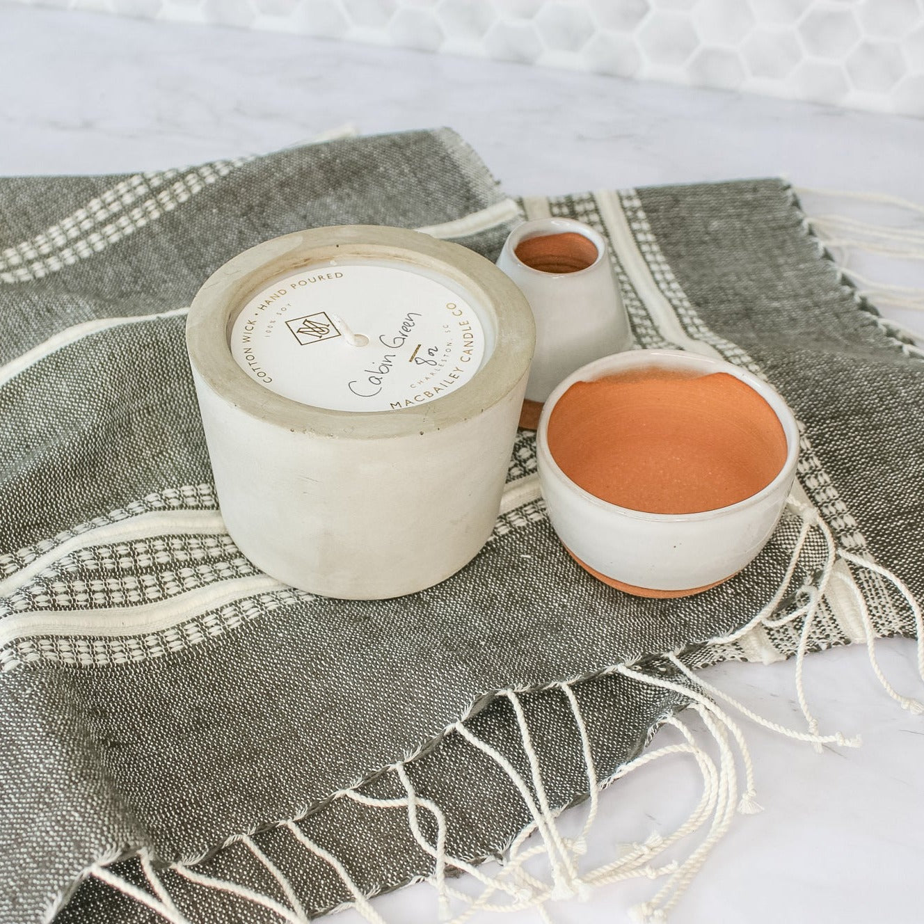 On a marble countertop, sits our Aden cotton hand towel. A dark grey towel with natural colored stripes and fringe on the ends. On top of the towel is our concrete candle in Cabin Green scent and two of our small planters.