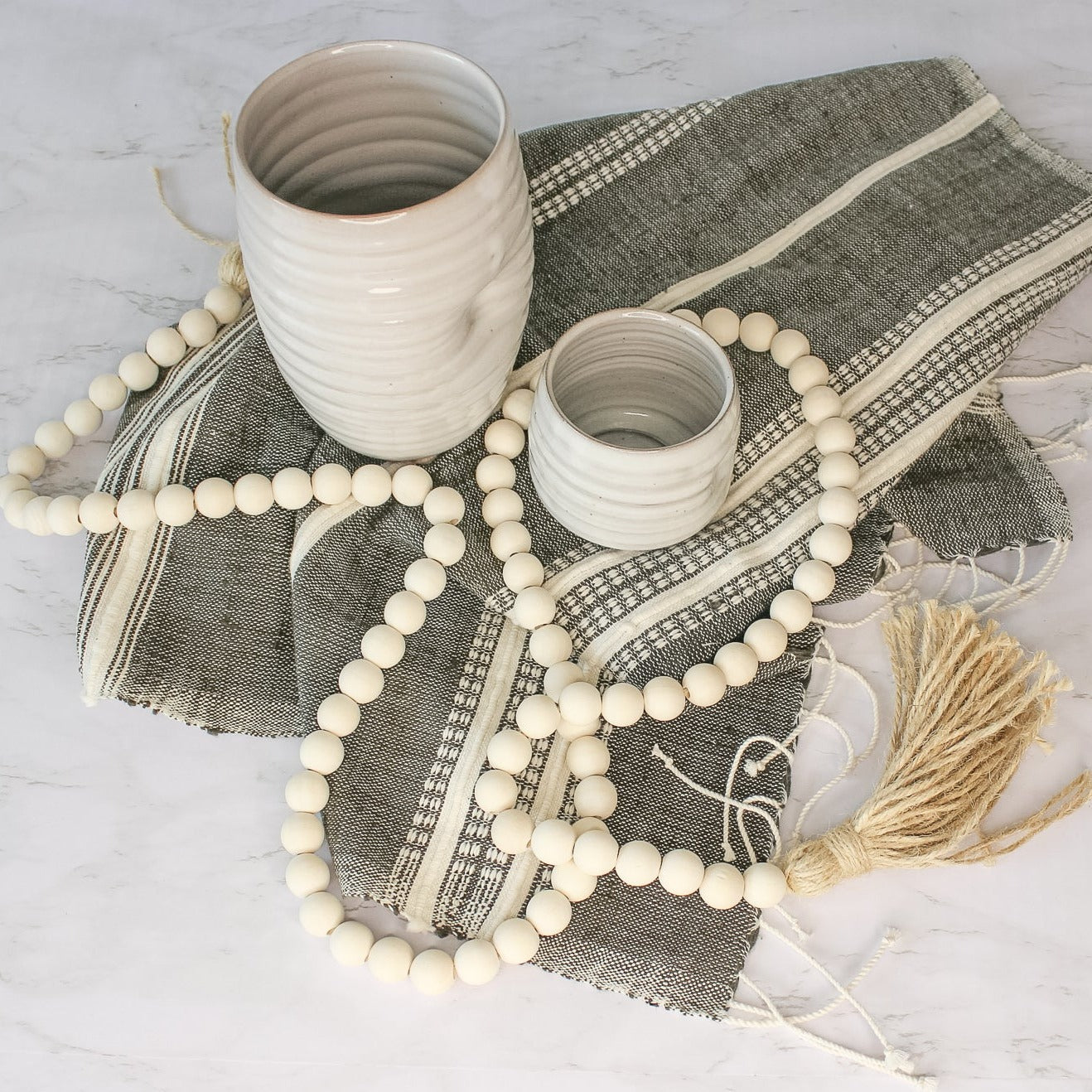 On a marble countertop, sits our aden cotton hand towel. A dark grey towel with natural colored stripes and fringe of the ends. On the towel, we have our tipsy tumbler and tall tumbler. Our decorative beads are draped over the towel. 