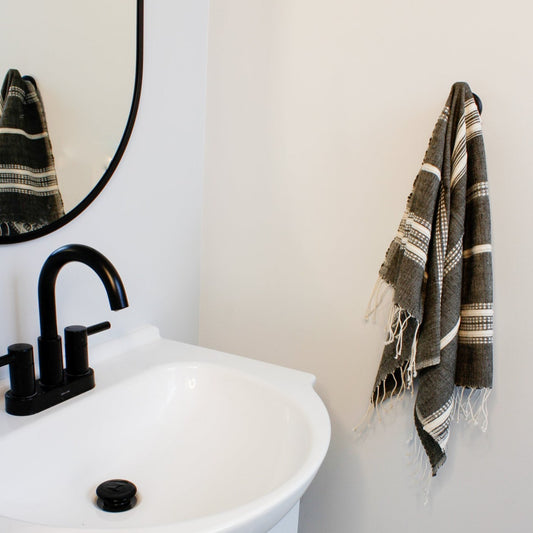 On a towel hook next to a sink, hangs our aden cotton hand towel. A dark grey towel with natural colored stripes and fringe on the ends.