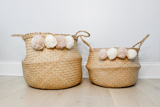 One large and one small natural belly baskets. Each have 4 pom poms along the edge. Pom poms are white and blush alternating colors.