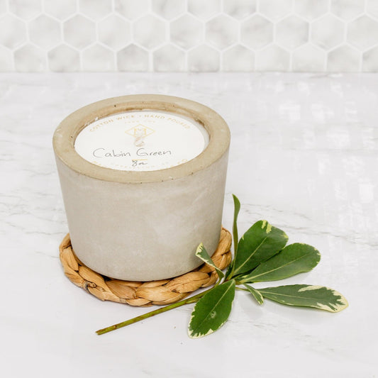 8oz concrete candle on seagrass coaster with greenery laying next to them. All resting on a marble counter top. 