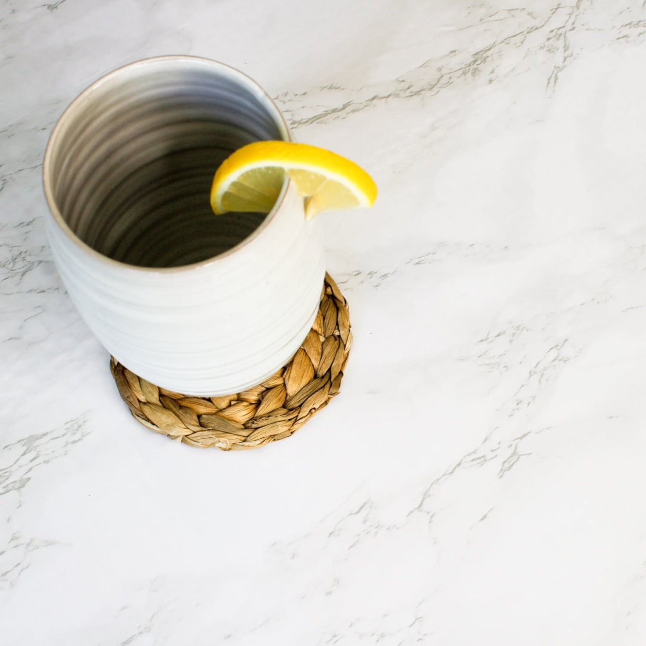 On a marble counter, sits our seagrass coaster and our tall tumbler with a lemon on the rim.
